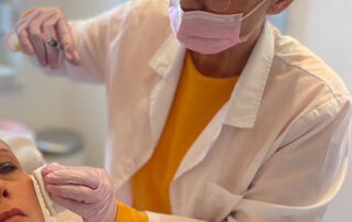 Ästhetische Behandlerin mit Maske und Handschuhen beugt sich leicht über eine Patientin, welche im Behandlungsstuhl liegt.