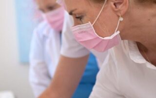 Heilpraktikerin in weißem Kittel, rosafarbenem Mundschutz beugt sich über einen Patientin (die man nicht sieht) und zeigt 2 weiteren Personen, welche hinter ihr stehen die Behandlung.